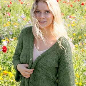 May include: A chunky knit olive green cardigan with a V-neckline and long sleeves. The garment features a loose, textured stitch pattern and is worn open over a light-colored base layer, showcasing a relaxed fit and a soft, slightly translucent appearance.