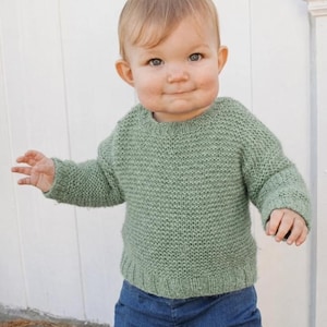 Puede incluir: Un suéter de punto de manga larga en color verde salvia con cuello redondo y puños y dobladillo acanalados, combinado con pantalones de mezclilla azules. El suéter presenta un patrón de tejido texturizado y uniforme en toda la prenda.