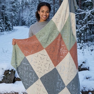 Puede incluir: Una colorida manta de punto con diseño de copo de nieve. La manta está hecha de cuadrados en tonos crema, verde, gris y óxido. La manta se muestra frente a un fondo de bosque nevado.