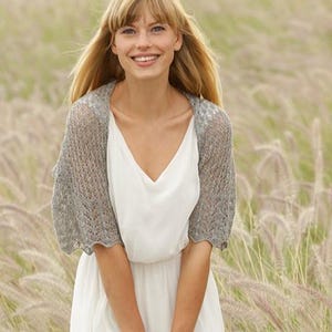 Può includere: Un coprispalle in maglia grigio chiaro aperto sul davanti, caratterizzato da un delicato e intricato motivo in pizzo con bordi smerlati e maniche corte, indossato sopra un semplice abito bianco senza maniche con scollo a V e tessuto fluido.