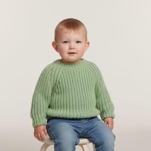 Peut inclure: Un enfant porte un pull en tricot vert clair et un jean bleu, assis sur un petit tabouret. Le pull a une texture côtelée et des manches longues. Le fond est blanc uni.
