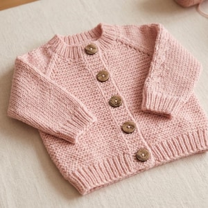 May include: A light pink knitted cardigan featuring a textured stitch pattern, long sleeves with ribbed cuffs, a ribbed crew neckline, and five round, dark brown wooden buttons down the front placket. The garment has a consistent knit texture.
