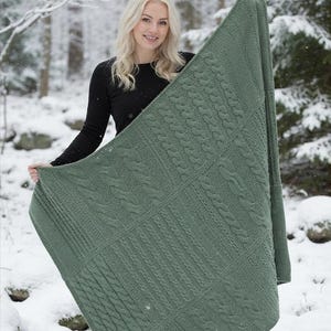Könnte beinhalten: Eine salbeigrüne Strickdecke mit einem strukturierten Zopfmuster, das aus abwechselnden vertikalen Bahnen mit geflochtenen Zöpfen und Rippstrick besteht. Die rechteckige Decke wird hochgehalten, um das komplexe Strickmuster zu zeigen.
