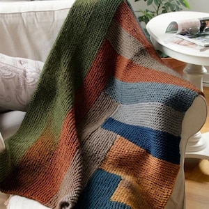 Könnte beinhalten: Eine handgestrickte Decke im Patchwork-Design. Die Decke hat ein Farbmuster mit Farbtönen von Olivgrün, Rostorange, Grau, Blau und Beige. Die Decke ist über ein beiges Sofa drapiert und zeigt ihre Textur und ihr Design.