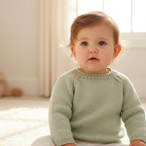 May include: A baby wearing a pale green knitted jumper, sitting on a cream carpet. The jumper has long sleeves and a round neck. The baby is looking at the camera. Wooden blocks are in the foreground.