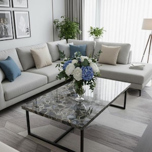 Könnte beinhalten: Ein modernes Wohnzimmer mit einem hellgrauen Ecksofa mit blauen und beigen Kissen. Ein rechteckiger Couchtisch mit gemusterter Oberfläche und schwarzem Metallrahmen steht auf einem gemusterten Teppich. Eine Vase mit Blumen sorgt für Farbe.