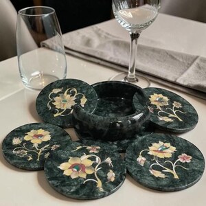 Könnte beinhalten: Ein Set aus runden Untersetzern aus dunkelgrünem Marmor mit eingelegten Blumenmustern in Gelb, Weiß und Rot. Die Untersetzer sind um einen passenden, zylindrischen Halter aus poliertem Marmor mit sichtbarer Steinmaserung angeordnet.