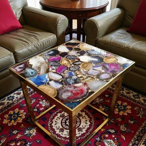 May include: Square coffee table with a mosaic top made of polished agate stones in shades of red, purple, blue, and brown. The table has a gold-colored metal frame and sits on a patterned red rug.