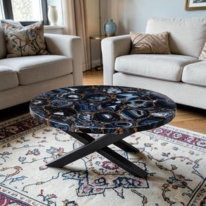 Könnte beinhalten: Ovaler Couchtisch mit einer schwarzen und blauen Achatsteinplatte und einem schwarzen Metallgestell in X-Form. Die Steinplatte hat ein einzigartiges Muster mit goldfarbenen Akzenten. Der Tisch steht auf einem gemusterten Teppich in einem Wohnzimmer.