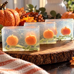 May include: Three rectangular bars of clear soap, each containing an embedded orange pumpkin shape with green vine details. The soap bars are arranged in a row, showcasing a translucent texture with small internal bubbles throughout the base.