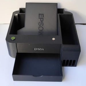May include: A black Epson printer with an open paper tray. The printer has the Epson logo on the front and the word Epson vertically on the top. A green power button is visible on the left side.