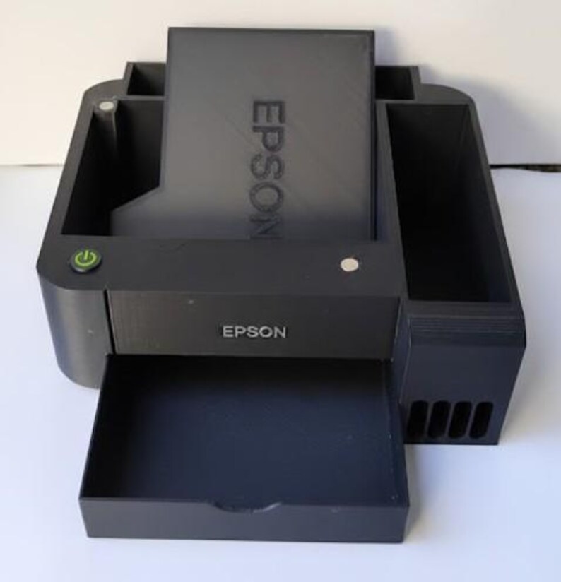May include: A black Epson printer with an open paper tray. The printer has the Epson logo on the front and the word Epson vertically on the top. A green power button is visible on the left side.