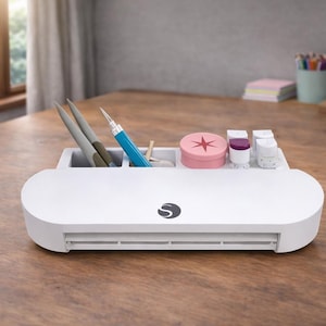 May include: White craft machine with a built-in storage compartment. The compartment holds pens, a blue craft tool, and small containers. A pink container with a star design and small white containers are also visible. The machine has a curved design and a logo.