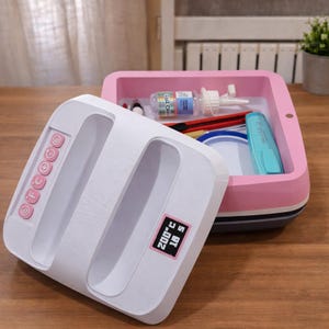 Puede incluir: Una plancha térmica blanca con botones rosas y una pantalla digital que muestra 200°C y 18 segundos. Un contenedor de almacenamiento rosa y blanco contiene suministros de manualidades, incluyendo pegamento, tijeras y un marcador azul. Sobre una superficie de madera.