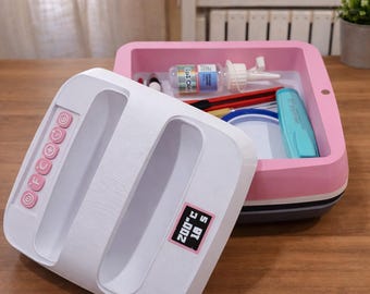 May include: A white heat press with pink buttons and a digital display showing 200°C and 18 seconds. A pink and white storage container holds craft supplies, including glue, scissors, and a blue marker. The items are on a wooden surface.