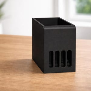 May include: A black desk organizer with a rectangular shape and a slanted top. The organizer has four vertical slots on the front and sits on a wooden surface. A second black object is visible to the left.