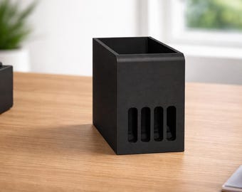 May include: A black desk organizer with a rectangular shape and a slanted top. The organizer has four vertical slots on the front and sits on a wooden surface. A second black object is visible to the left.
