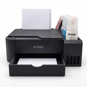 May include: A black Epson printer with a paper tray and ink bottles. The printer has a stack of white paper in the input tray. The Epson logo is visible on the front of the printer and on the ink bottles.
