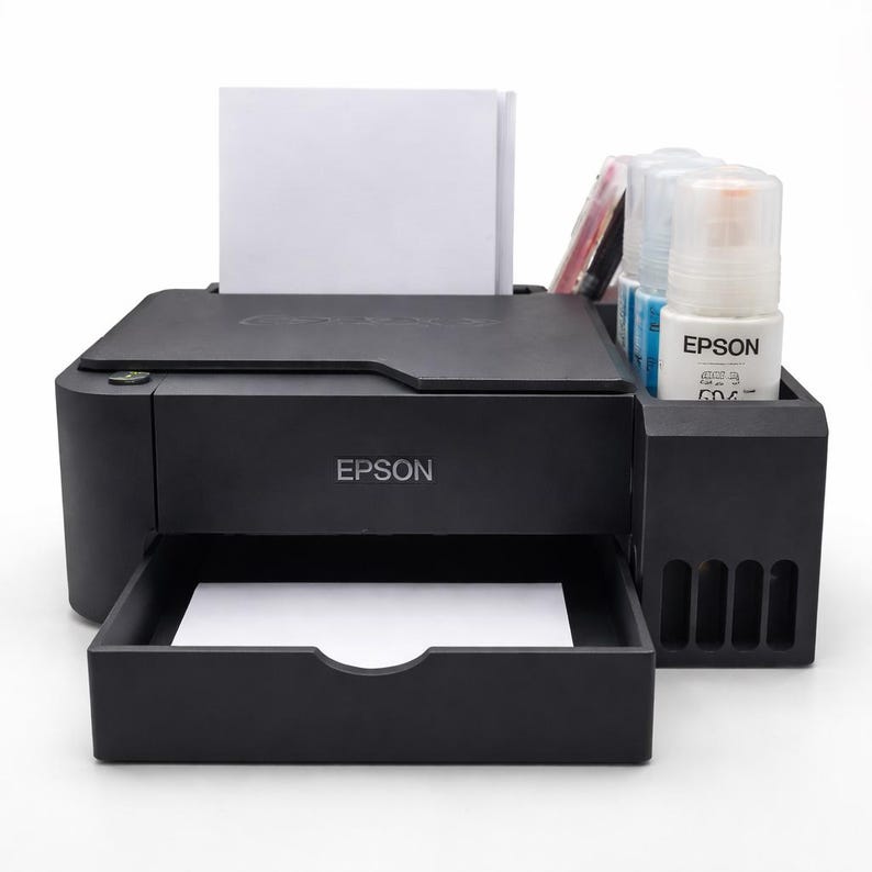 May include: A black Epson printer with a paper tray and ink bottles. The printer has a stack of white paper in the input tray. The Epson logo is visible on the front of the printer and on the ink bottles.
