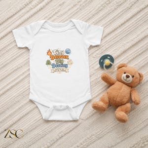 May include: A white short-sleeved baby bodysuit featuring a graphic with the text Tiny Bender, Big Destiny, mastering naps, milk & all 4 elements, surrounded by icons of fire, water, earth, and air, accompanied by a blue pacifier and a brown teddy bear.