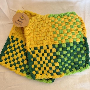 Op de afbeelding: Twee geweven pannenlappen in geel en groen. De pads hebben een dambordpatroon. De tekst "Hayes Handy Hot Pads" staat op een klein label dat aan een van de pads is bevestigd.