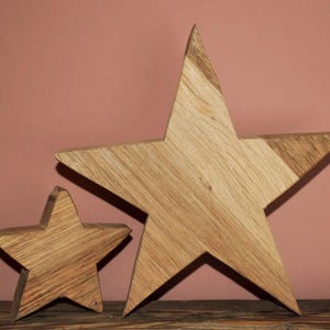 Könnte beinhalten: Zwei fünfzackige Sterne aus hellem Holz mit sichtbarer Maserung. Der größere Stern ist rechts positioniert, während ein kleinerer Stern links platziert ist. Beide Objekte haben eine flache, glatte Oberfläche und natürliche Holztöne.