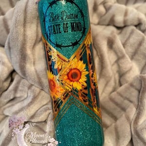 May include: A teal glitter tumbler with a sunflower and leopard print design. The tumbler has the text "Beth Dutton STATE OF MIND" in a black circle. The bottom of the tumbler has the Moon Blossom Creations logo.