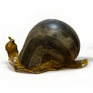 以下が含まれることがあります： 暗い茶色の殻と明るい茶色の体を持つセラミックカタツムリの彫刻。 殻はテクスチャ加工されたストライプのデザインです。 カタツムリの体は光沢のある明るい茶色で、白い背景に配置されています。