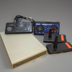May include: A collection of vintage video game consoles and controllers. Includes a beige Atari console, two black joysticks with orange accents, and two controllers, one black and one blue, with the text "TurboPad" and "Acclaim - Double Player".