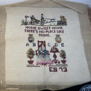 以下が含まれることがあります： 「HOME SWEET HOME THERE'S NO PLACE LIKE HOME」の文字が入った、ヴィンテージのクロスステッチタペストリー。馬車、椅子に座った女性、鉢植えの植物が描かれています。ベージュ、茶色、青色を基調とした色使いです。