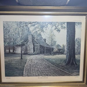 Op de afbeelding: Ingelijste prent van een huis op een heuvel, met een bakstenen pad dat leidt naar een blokhut met een stenen schoorsteen. Het kunstwerk is in grijstinten en heeft een goudkleurige lijst. Gesigneerd door Fred Thrasher.