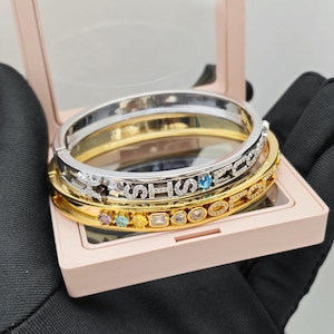 Puede incluir: Una pila de tres brazaletes dorados y plateados, uno con la palabra "SHOP" escrita con diamantes y piedras preciosas. Los brazaletes se exhiben en una caja de joyería rosa.