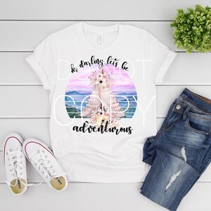 May include: White t-shirt with a colorful graphic featuring a floral design and the text "So darling let's be adventurous." The shirt is paired with blue jeans and white sneakers, set against a white wooden background.