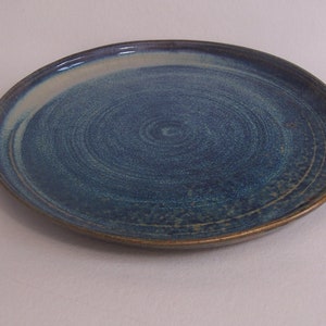 May include: A ceramic plate with a blue and teal glaze. The plate has an irregular shape and a spiral pattern in the centre. The rim is a darker brown colour. A unique piece for serving food, approximately 25 cm in diameter.
