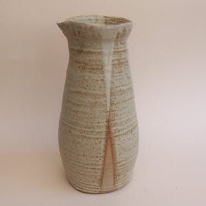 May include: A tall, tapered ceramic jug with a speckled off-white glaze and horizontal textured ridges. The surface features a vertical drip pattern in a muted brown tone that extends from the rim down to the base of the vessel.