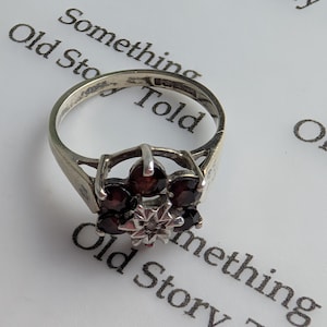 May include: A silver-toned ring featuring a cluster of six round, deep red garnet stones arranged in a floral pattern. At the centre of the garnet cluster sits a small, star-shaped metal setting holding a tiny, clear, faceted stone.