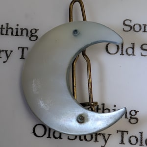 May include: A silver crescent moon hair clip with a gold-coloured metal clasp. The moon has a pearlescent finish and is attached to the clip with two small rivets. The clip is set against a white background with text.