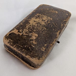 May include: A rectangular, hinged storage case with a distressed, dark brown textured exterior finish. The case features rounded corners and a small, thin metal latch protruding from the side, showing signs of age and wear on the surface.