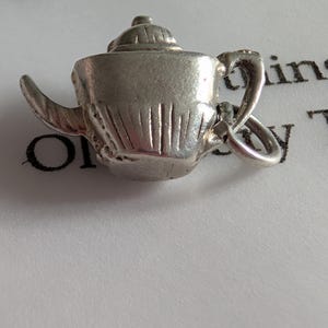 May include: A miniature silver teapot charm. The charm has a detailed design, including a spout, handle, and lid. It is attached to a small ring. The teapot is likely made of metal and is a decorative item, possibly for a bracelet or necklace.