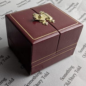 May include: A burgundy jewelry box with gold trim and a decorative gold clasp. The box is rectangular and has a hinged lid. The background is a patterned surface with the text "Something Old Story Told".