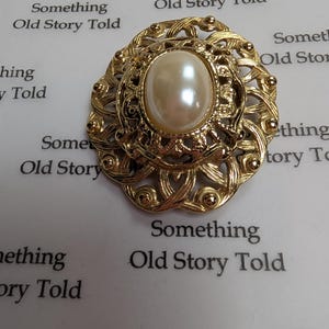 May include: A gold-coloured vintage brooch with a cream-coloured oval faux pearl at its centre. The brooch has an ornate, openwork design with intricate details and a textured finish. The text "Something Old Story Told" is visible.