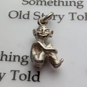 May include: A silver goblin charm, seated with its knees drawn up, featuring a smiling face and large ears. The charm is designed to be added to a bracelet or necklace. The background has text that reads "Something Old Story Told."