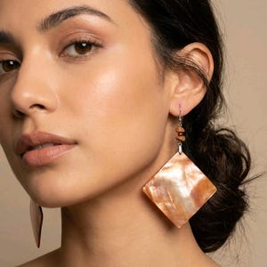 May include: Diamond-shaped earrings with an iridescent, peach-coloured shell design. The earrings have small, brown beads and silver-coloured hooks. A statement piece of jewellery.