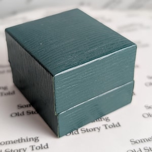 May include: A small, square, teal-colored box with a textured, ribbed surface. The box is closed, with a visible seam where the lid meets the base. The background features printed text that reads "Something Old Story Told".