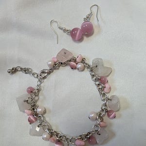May include: A silver-toned charm bracelet and a pair of matching drop earrings. The jewellery set features pink cat's eye beads, small white pearls, and translucent pale pink square-cut stones attached to a silver-toned metal chain link bracelet.