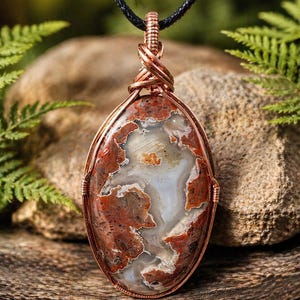May include: Oval pendant featuring a polished stone with red, white, and brown patterns, encased in copper wire. The pendant hangs from a black cord, showcasing a natural, earthy aesthetic. The stone's unique patterns create a striking visual appeal.
