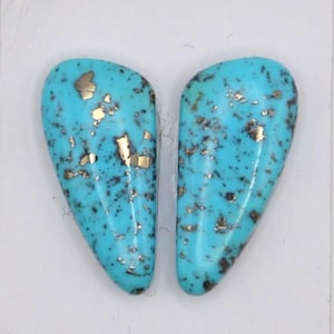 May include: Two turquoise gemstone cabochons, teardrop-shaped, with gold-coloured inclusions and a speckled pattern. The background is white.