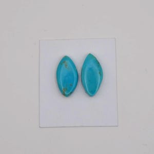 May include: Two marquise-cut turquoise gemstones. The stones are a vibrant blue-green colour with natural brown veining. They are set on a white surface, showcasing their polished, smooth surfaces. These gemstones are ideal for jewellery making.