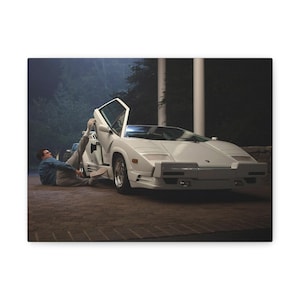 May include: A white Lamborghini Countach sports car with its distinctive scissor door open. The car is parked on a brick surface, with a person lying next to it. The scene is set at night, with trees and a building in the background.