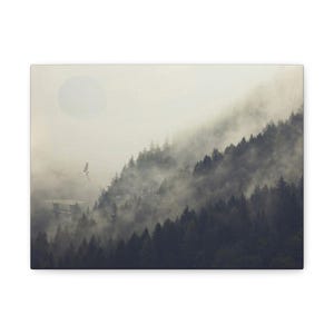 May include: A canvas print featuring a misty mountain landscape with a forest of dark green trees. A large, pale orb is in the sky, and two flying objects are visible. The scene is shrouded in fog, creating a serene and atmospheric artwork.